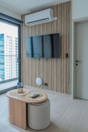 Ein modernes Hotelzimmer mit großem Fenster, einem an einer Holzvertäfelung befestigten Flachbildfernseher, einem runden Tisch und einer Fernbedienung sowie einer Tür rechts.