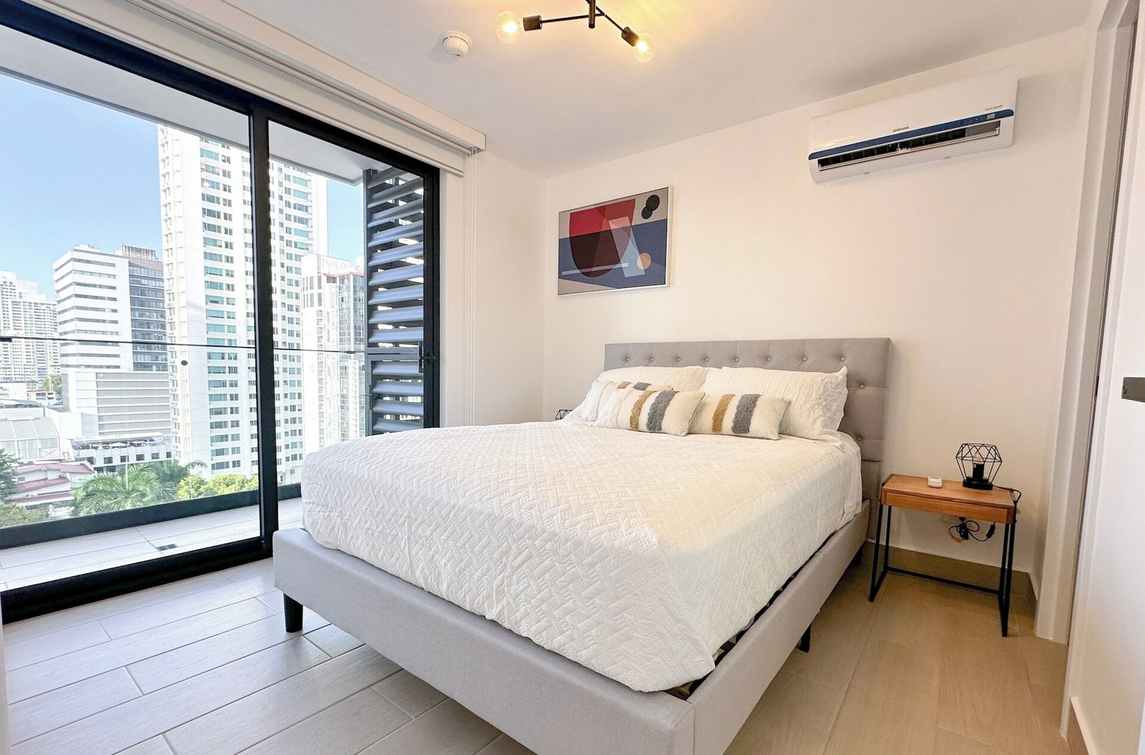 A modern bedroom with a large bed, a nightstand, a wall-mounted air conditioner, and a city view.