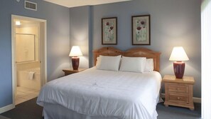 A hotel room with a bed, two bedside tables with lamps, a door, and two wall-mounted paintings. - Waterside by Spinnaker - 2BR Hilton Head Condo, Short Walk to the Beach (Hilton Head Island)
