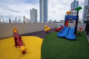 Ein Dachgarten mit Spielplatz, inklusive Wippe, Rutsche und Klettergerüst, umgeben von Hochhäusern.