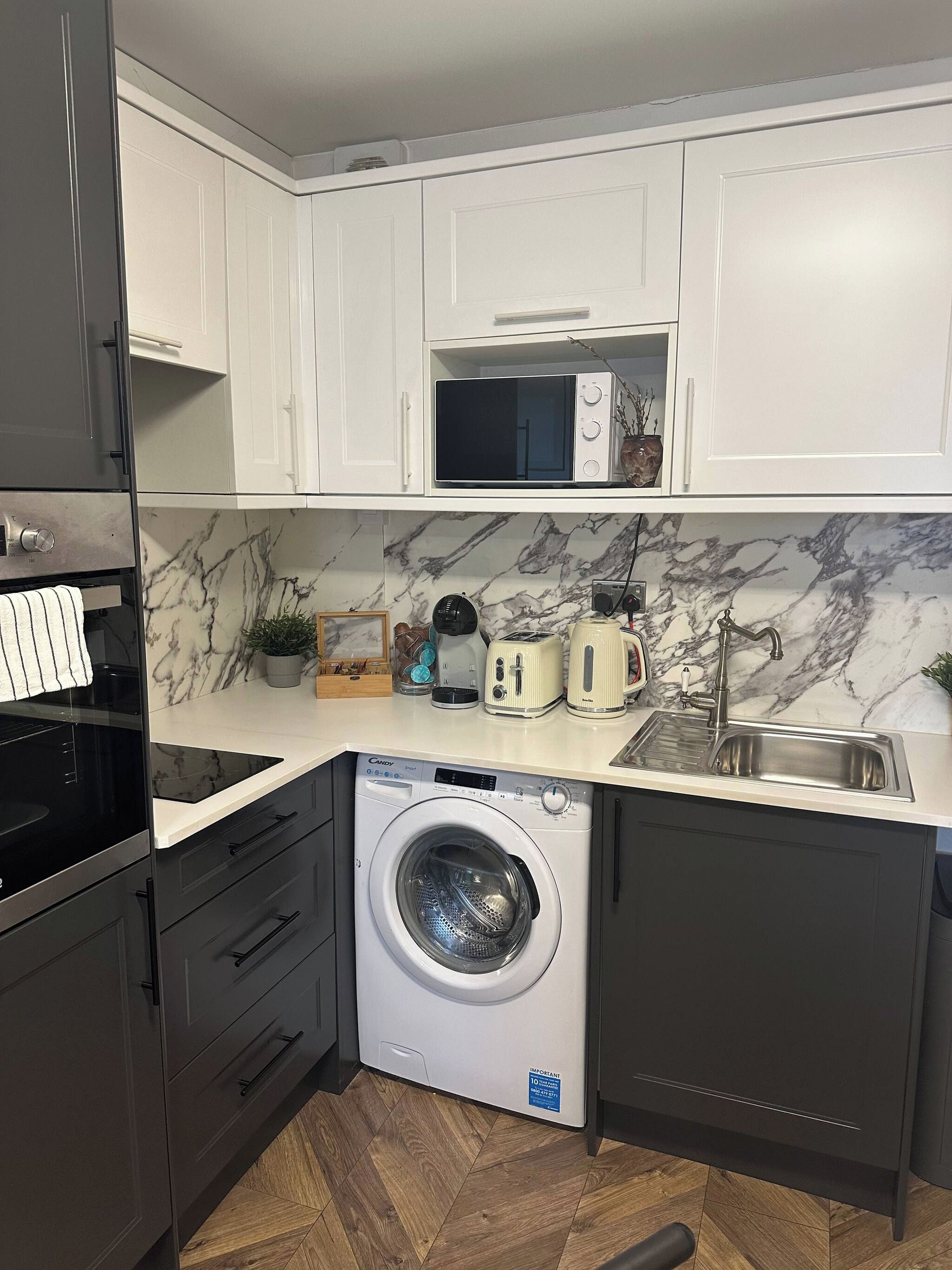 A modern kitchen with white cabinets, a built-in microwave, a washing machine, and a sink.