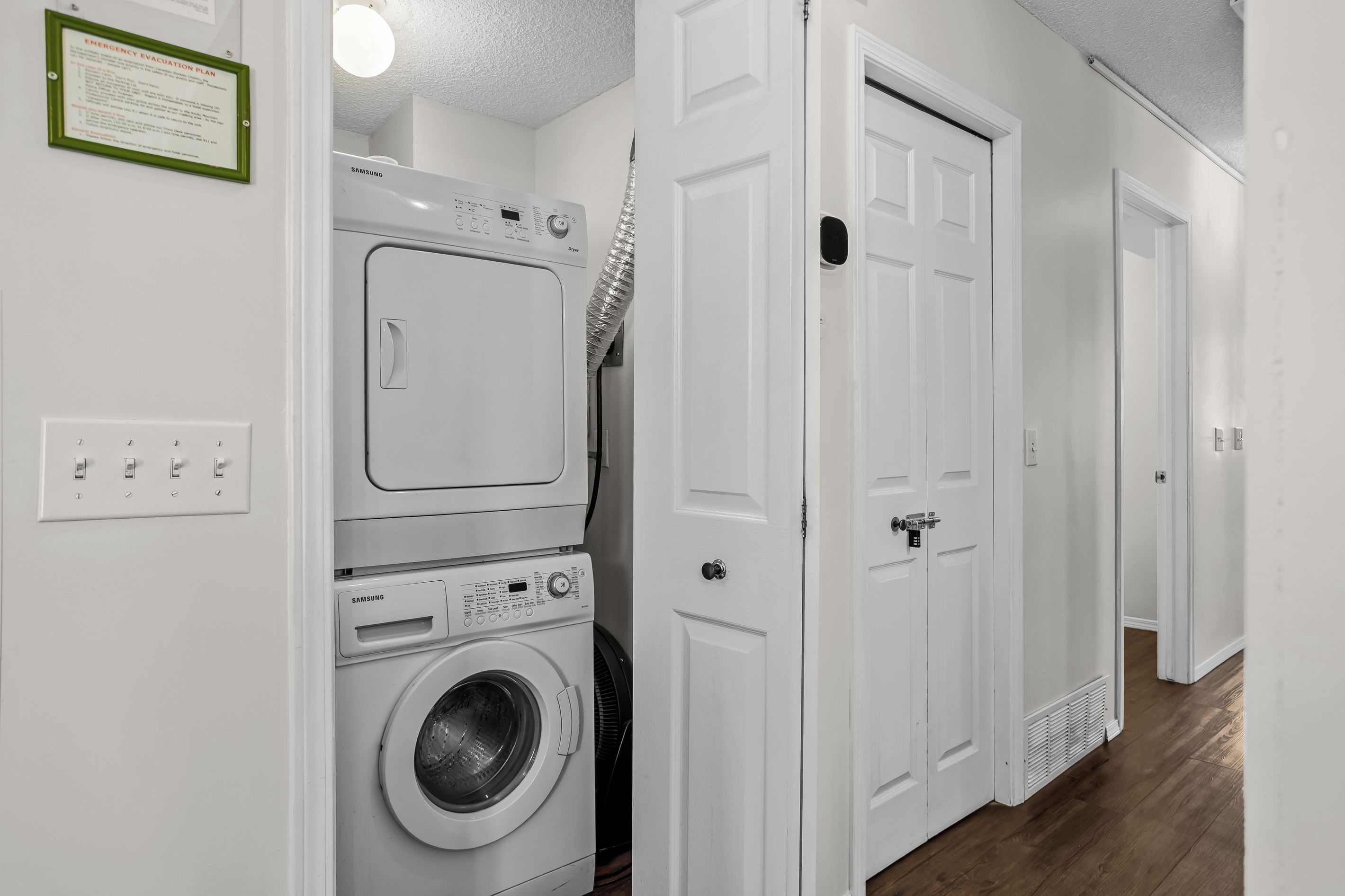 Une buanderie équipée d’un lave-linge et d’un sèche-linge superposés, d’une porte dotée d’un judas et d’un luminaire fixé au mur.