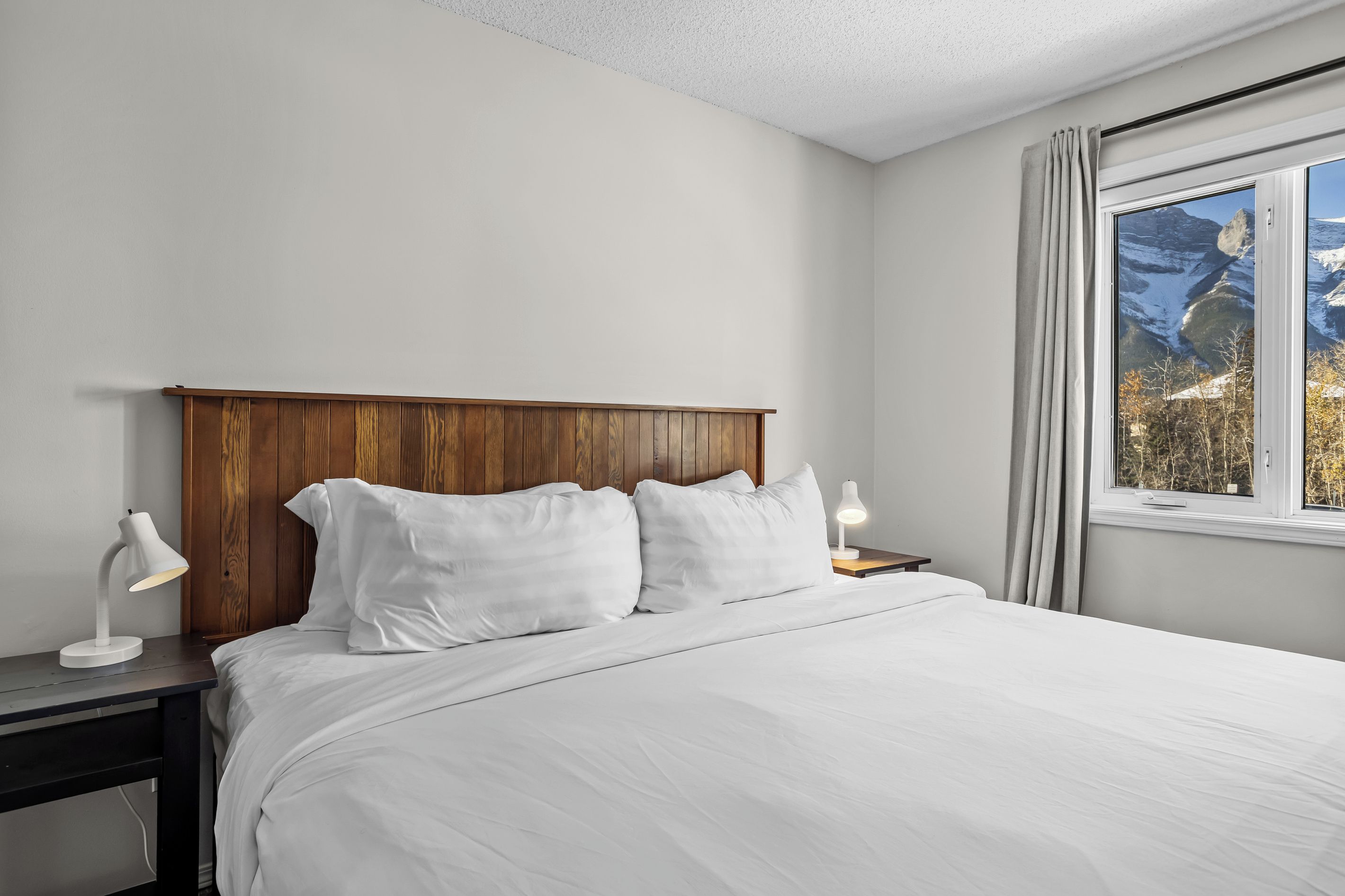 A bedroom with a wooden headboard, white bedding, a desk with a lamp, and a window with a view of mountains.