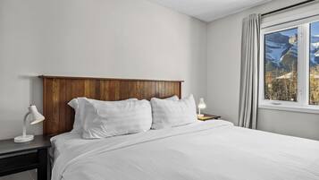 A bedroom with a wooden headboard, white bedding, a desk with a lamp, and a window with a view of mountains.