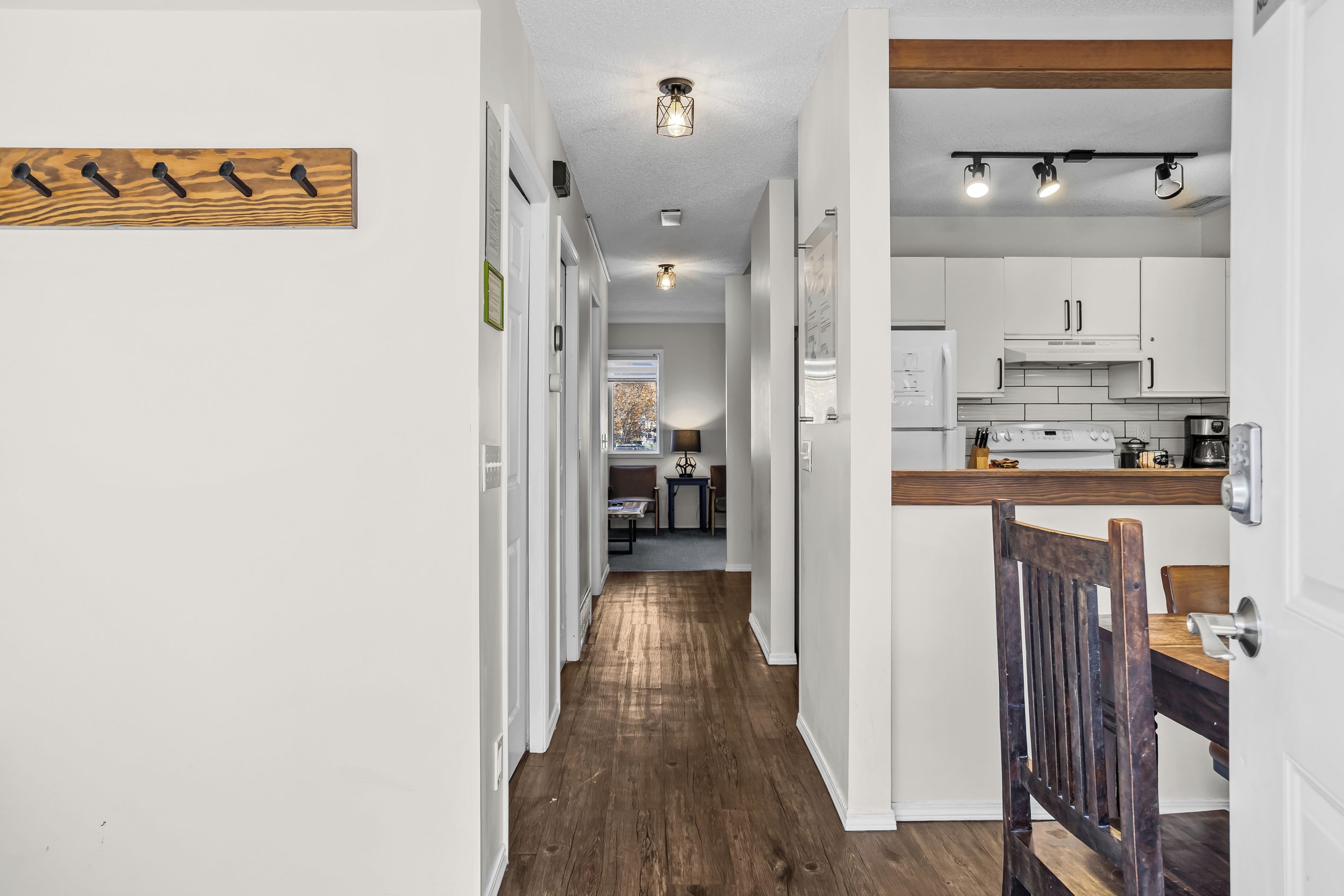 A hallway leading to a kitchen with white cabinets, a wooden floor, and a dining area with a wooden chair.