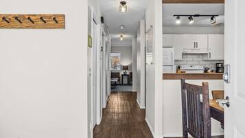 A hallway leading to a kitchen with white cabinets, a wooden floor, and a dining area with a wooden chair.