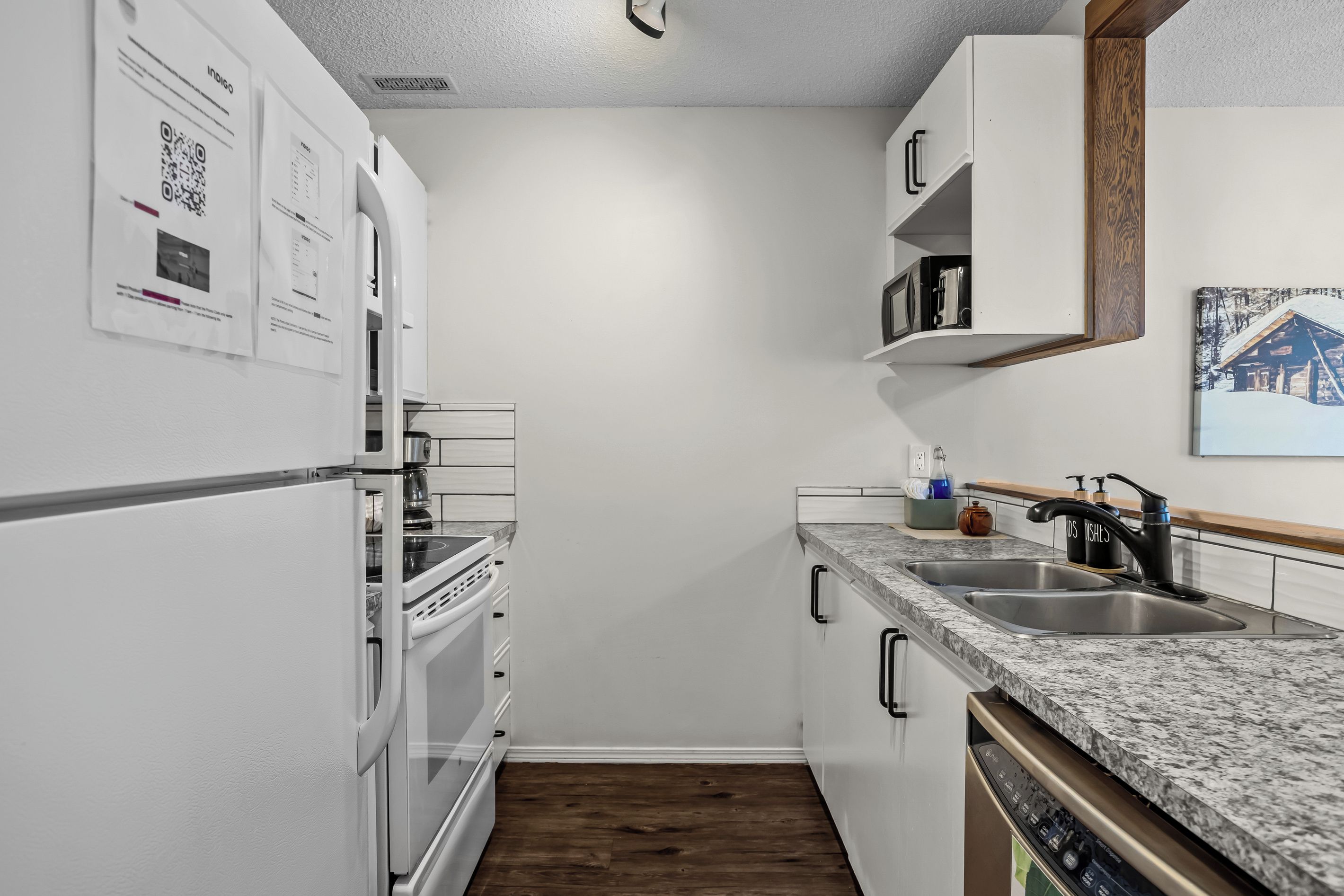 Ett kompakt kök med vita skåp, en inbyggd ugn, en diskmaskin och en diskho med en modern blandare.