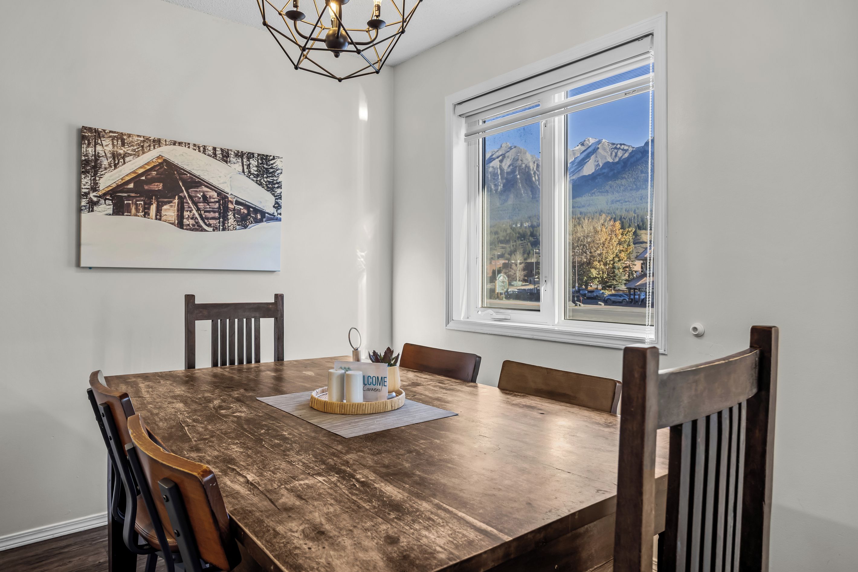 Un coin repas avec une table en bois, des chaises, un lustre, une fenêtre donnant sur la montagne et un tableau encadré au mur.