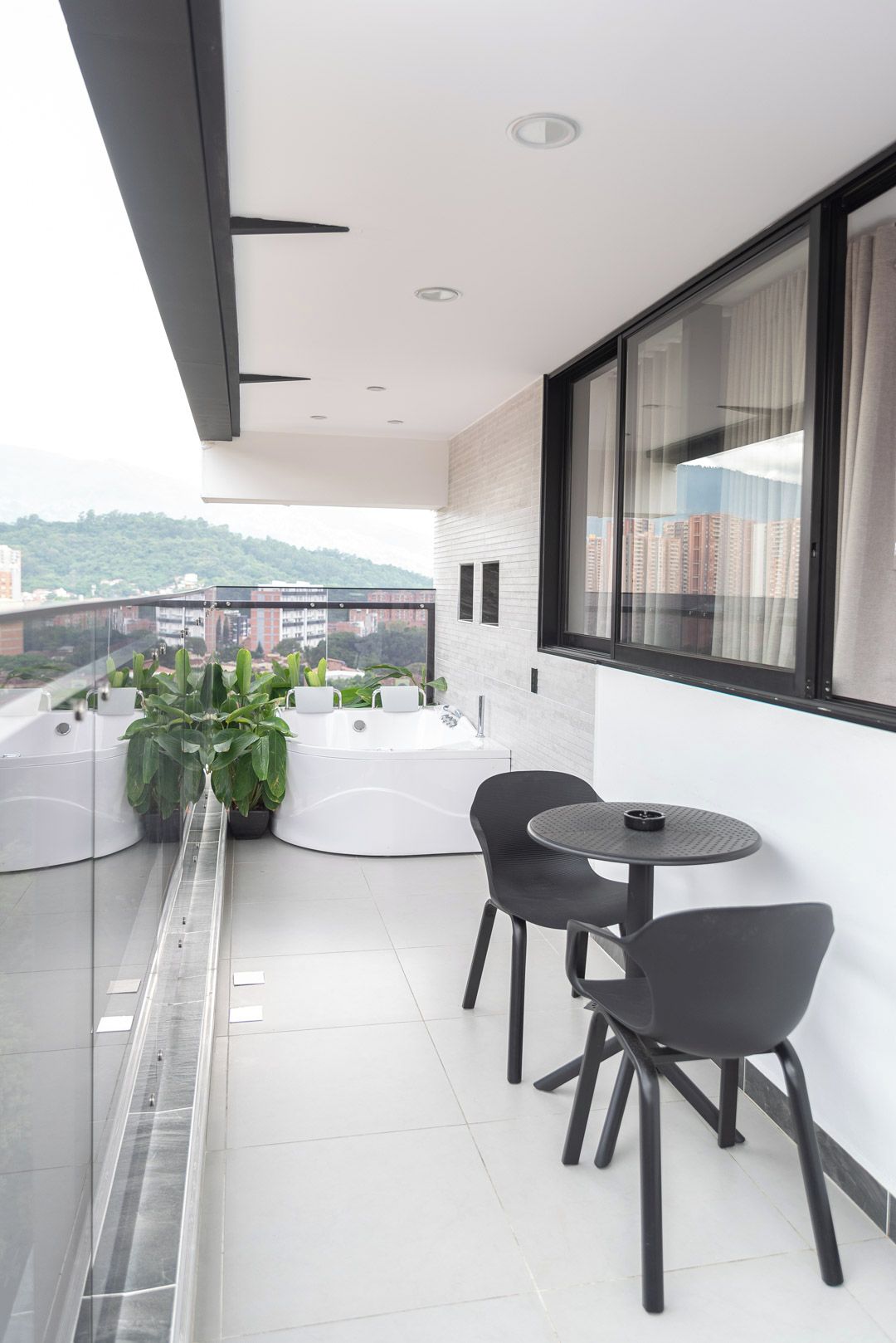 En moderne balkon med glasværn, to stole, et lille rundt bord og udsigt over bybilledet.