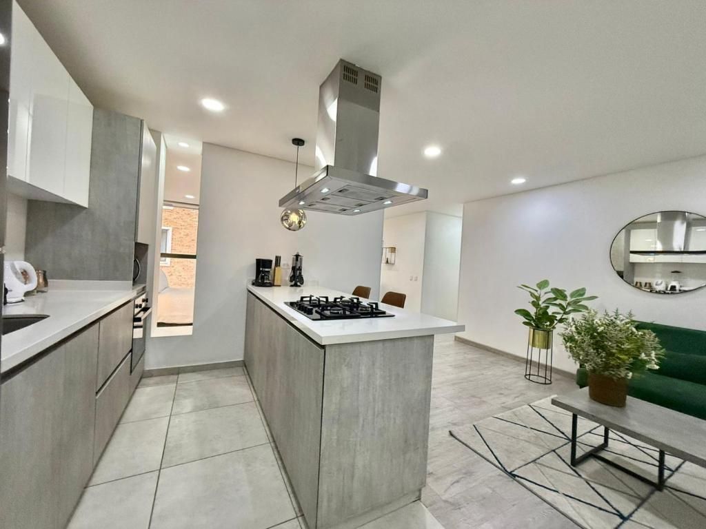 Una cocina moderna con una isla central, una estufa y una campana extractora. Hay un área de comedor con un sofá verde y un espejo redondo en la pared.
