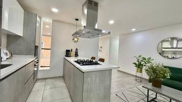Una cocina moderna con una isla central, una estufa y una campana extractora. Hay un área de comedor con un sofá verde y un espejo redondo en la pared.