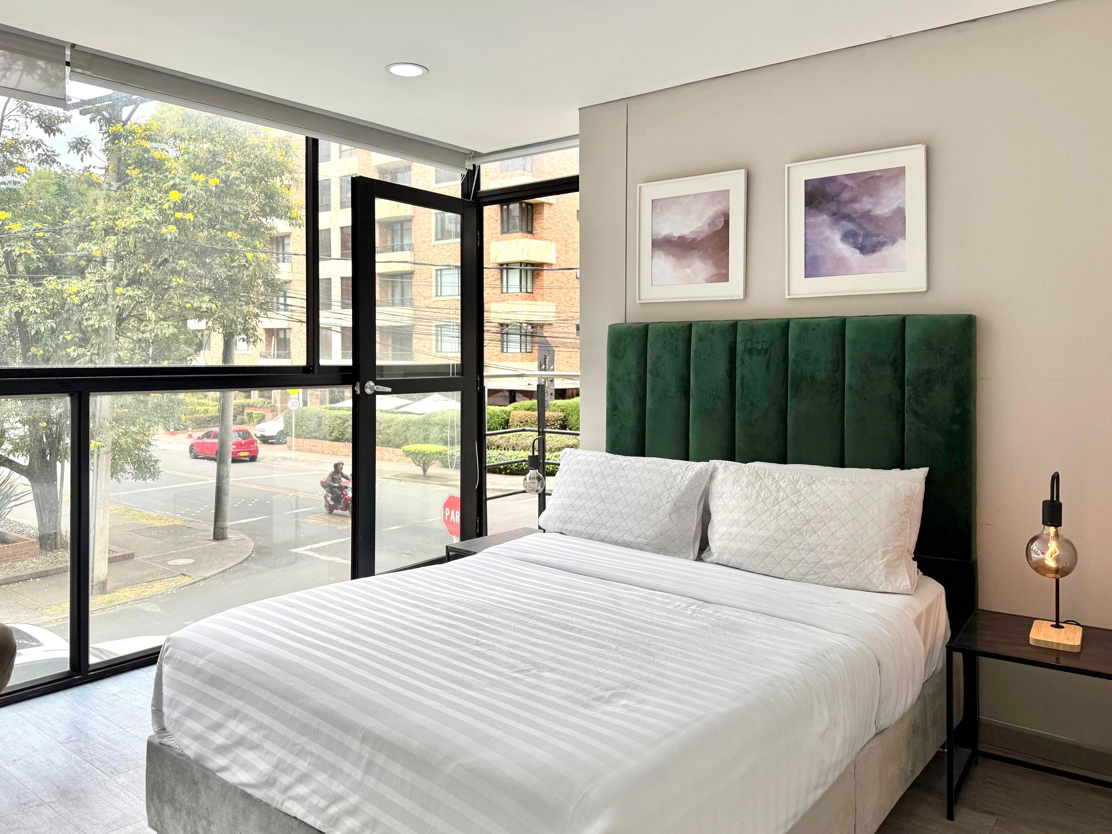 Un dormitorio moderno con un ventanal grande, un cabecero tapizado en verde, una colcha blanca y dos obras de arte enmarcadas en la pared.