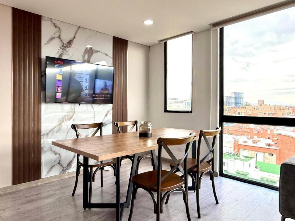 Un comedor moderno con una mesa de madera y sillas metálicas, un televisor montado en la pared, y un ventanal con vistas a la ciudad.