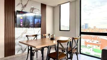 Un comedor moderno con una mesa de madera y sillas metálicas, un televisor montado en la pared, y un ventanal con vistas a la ciudad.