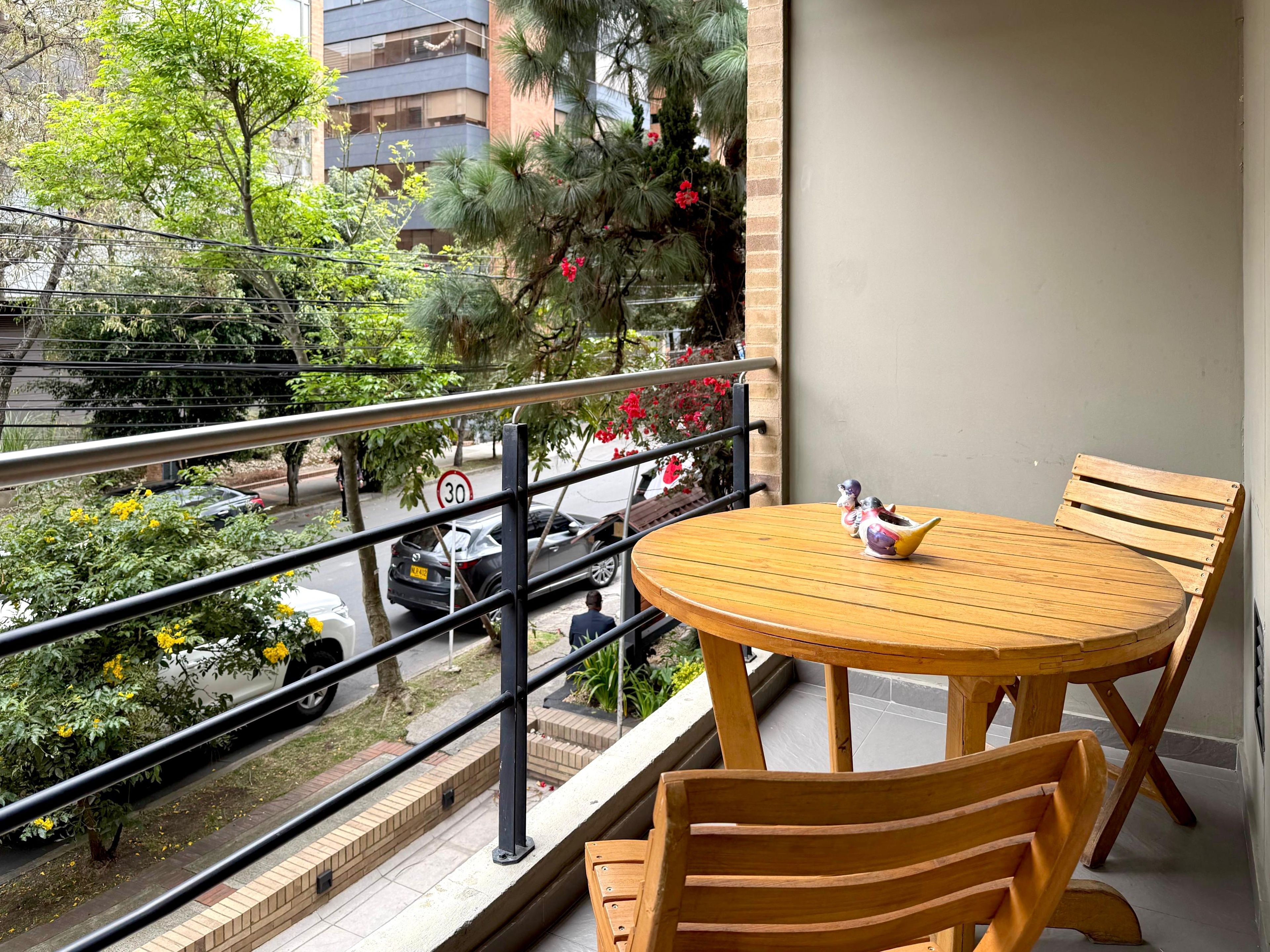 Un balcon avec une table ronde en bois et des chaises, une balustrade, et une vue sur la rue, avec des voitures et des arbres.