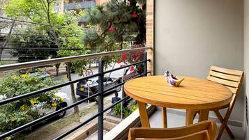 Un balcon avec une table ronde en bois et des chaises, une balustrade, et une vue sur la rue, avec des voitures et des arbres.
