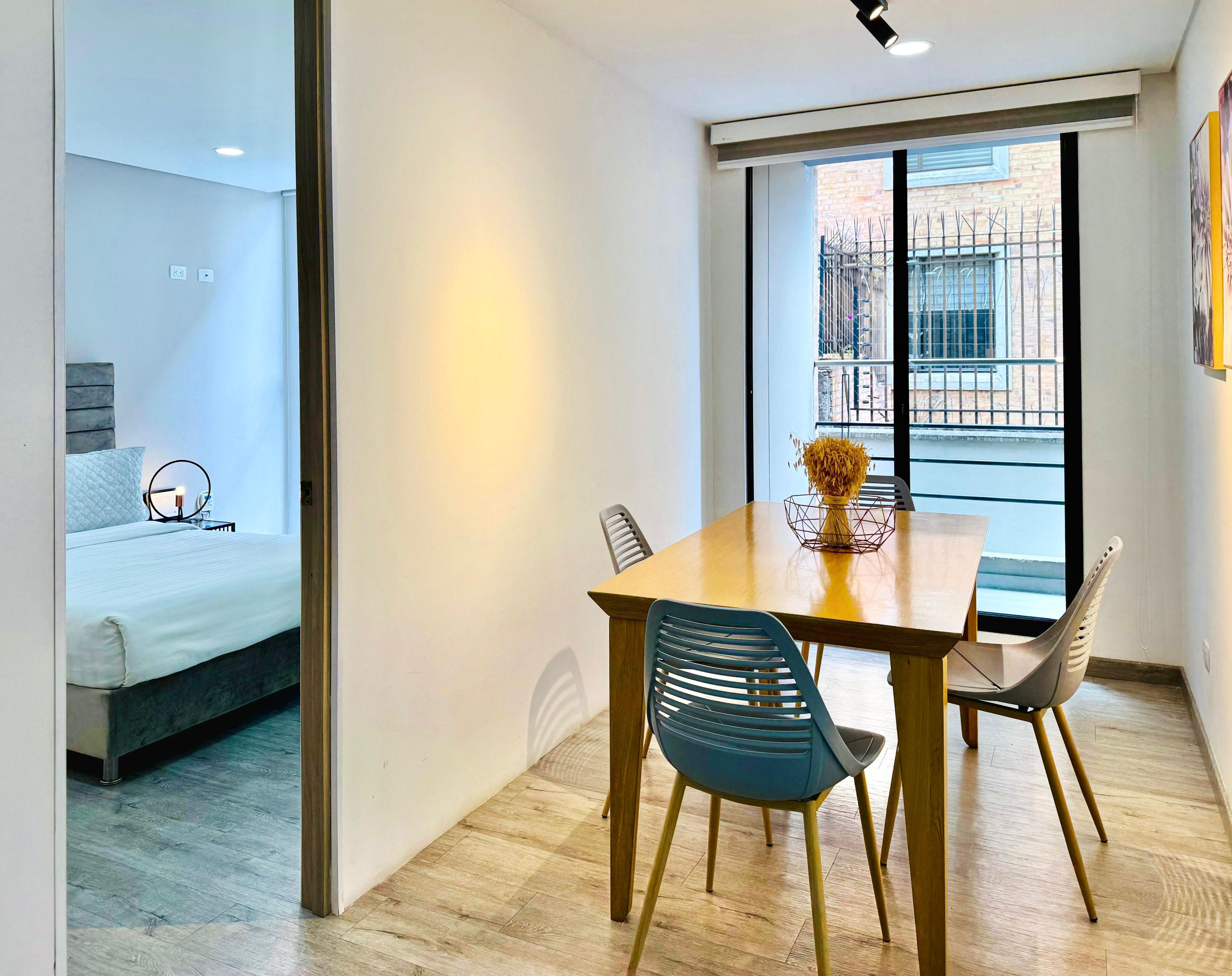 Une chambre d’hôtel moderne avec une table à manger en bois, deux chaises, un vase contenant des fleurs séchées et un lit avec une tête de lit grise.