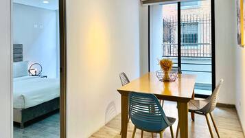 Une chambre d’hôtel moderne avec une table à manger en bois, deux chaises, un vase contenant des fleurs séchées et un lit avec une tête de lit grise.