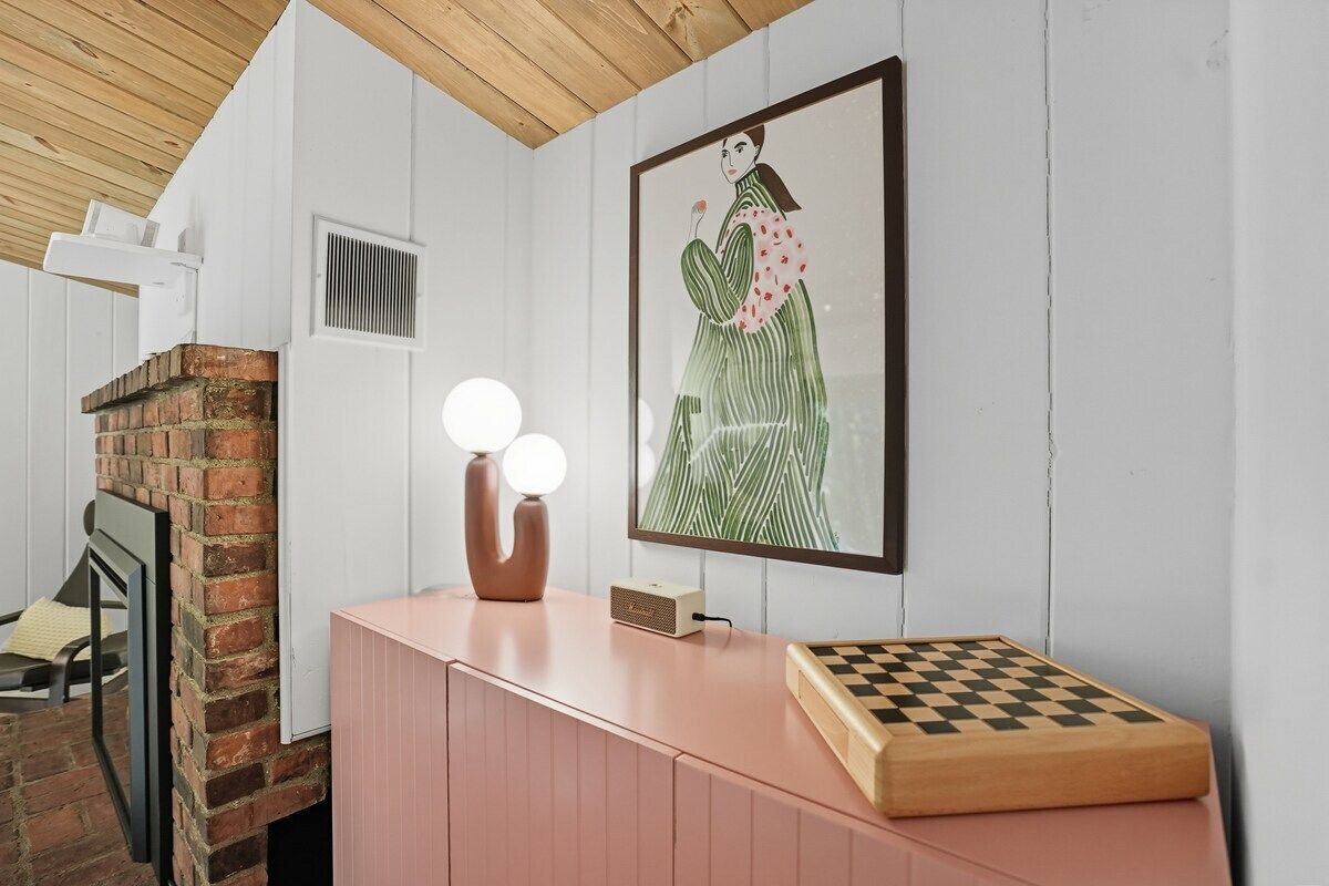 Una habitación con chimenea de ladrillo, un armario rosa, una obra de arte enmarcada y un tablero de ajedrez.
