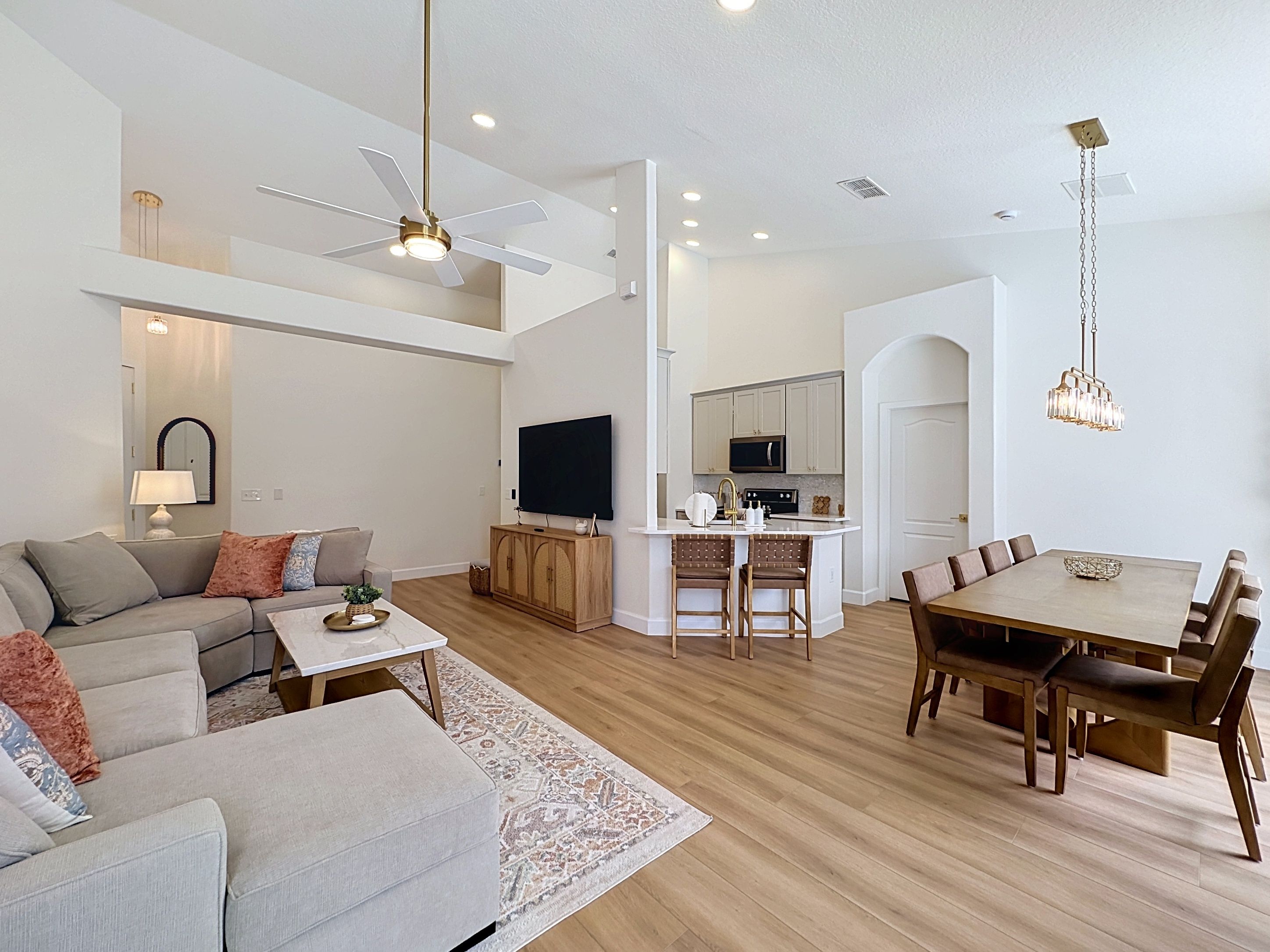 A modern living room with a sofa, coffee table, and a TV. There is a dining area with a table and chairs. The kitchen is visible with a microwave and cabinets.