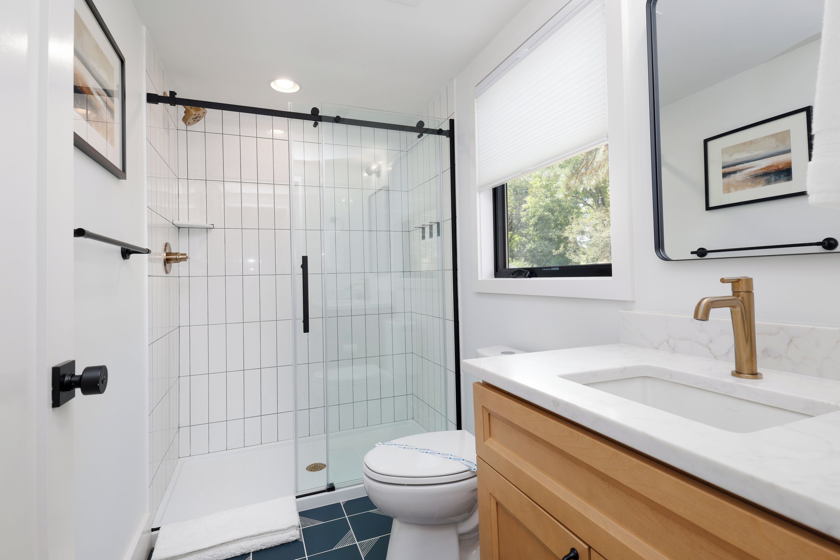 A modern bathroom with a glass shower enclosure, white tiles, a white toilet, a white sink with a gold faucet, and a wooden vanity.