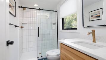 A modern bathroom with a glass shower enclosure, white tiles, a white toilet, a white sink with a gold faucet, and a wooden vanity.