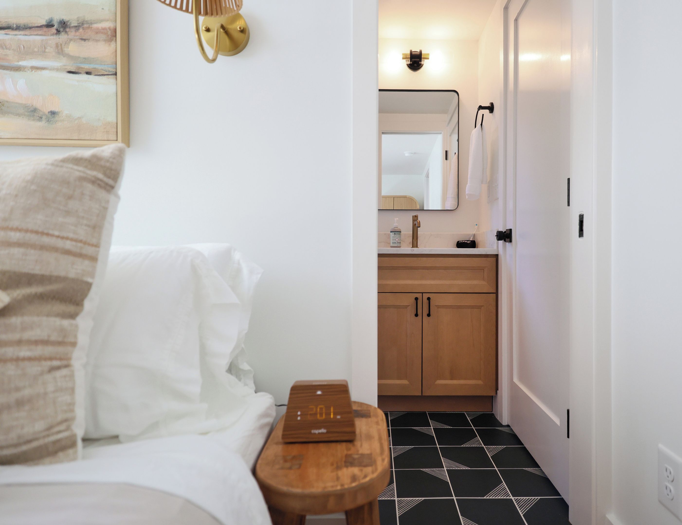 A hotel room with a bed, a bedside table with a clock, a small wooden stool, a painting on the wall, and a view into the bathroom with a mirror and sink.