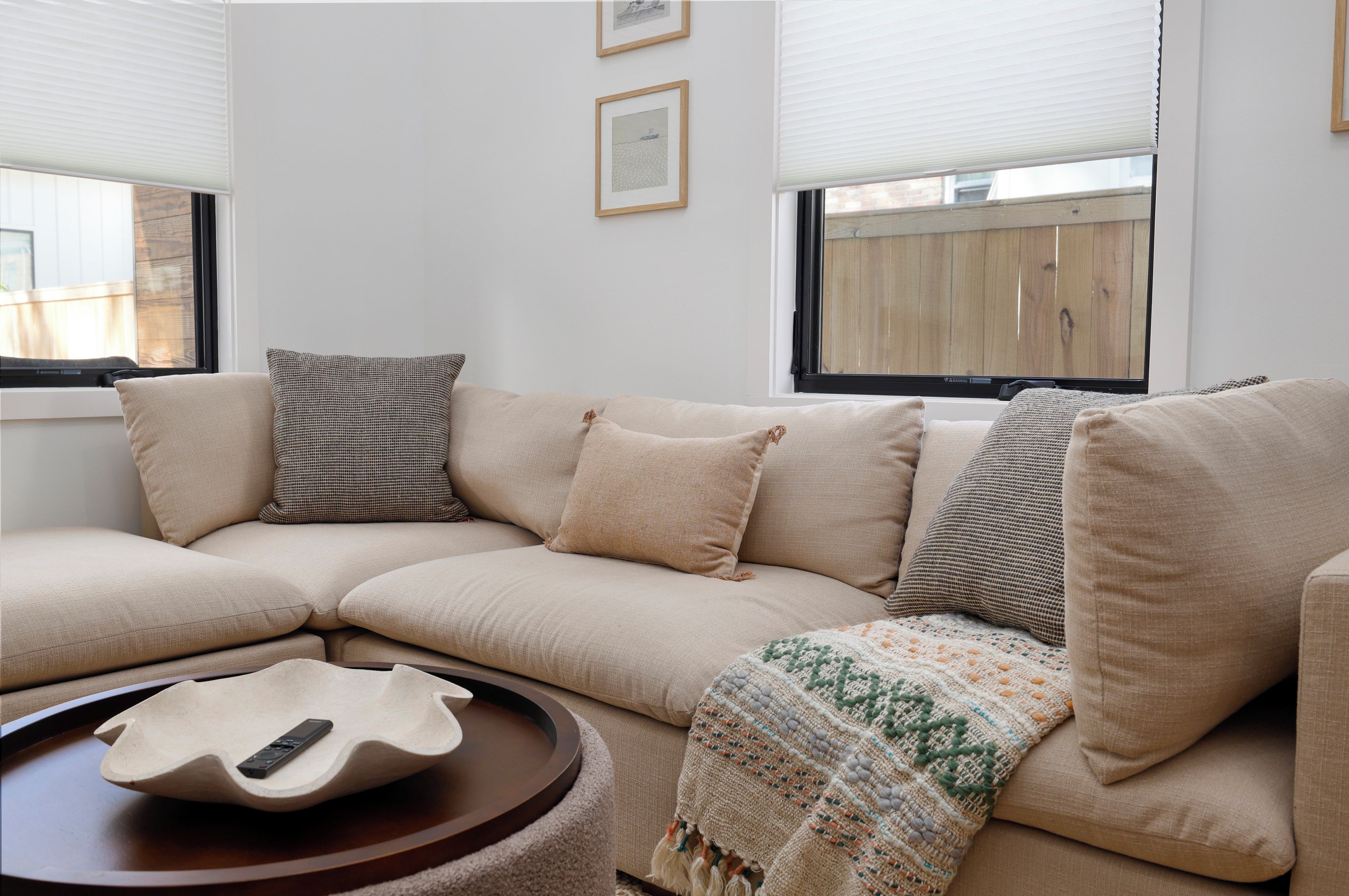 A living room with a beige sectional sofa, a remote control on a round wooden table, and a woven throw blanket.