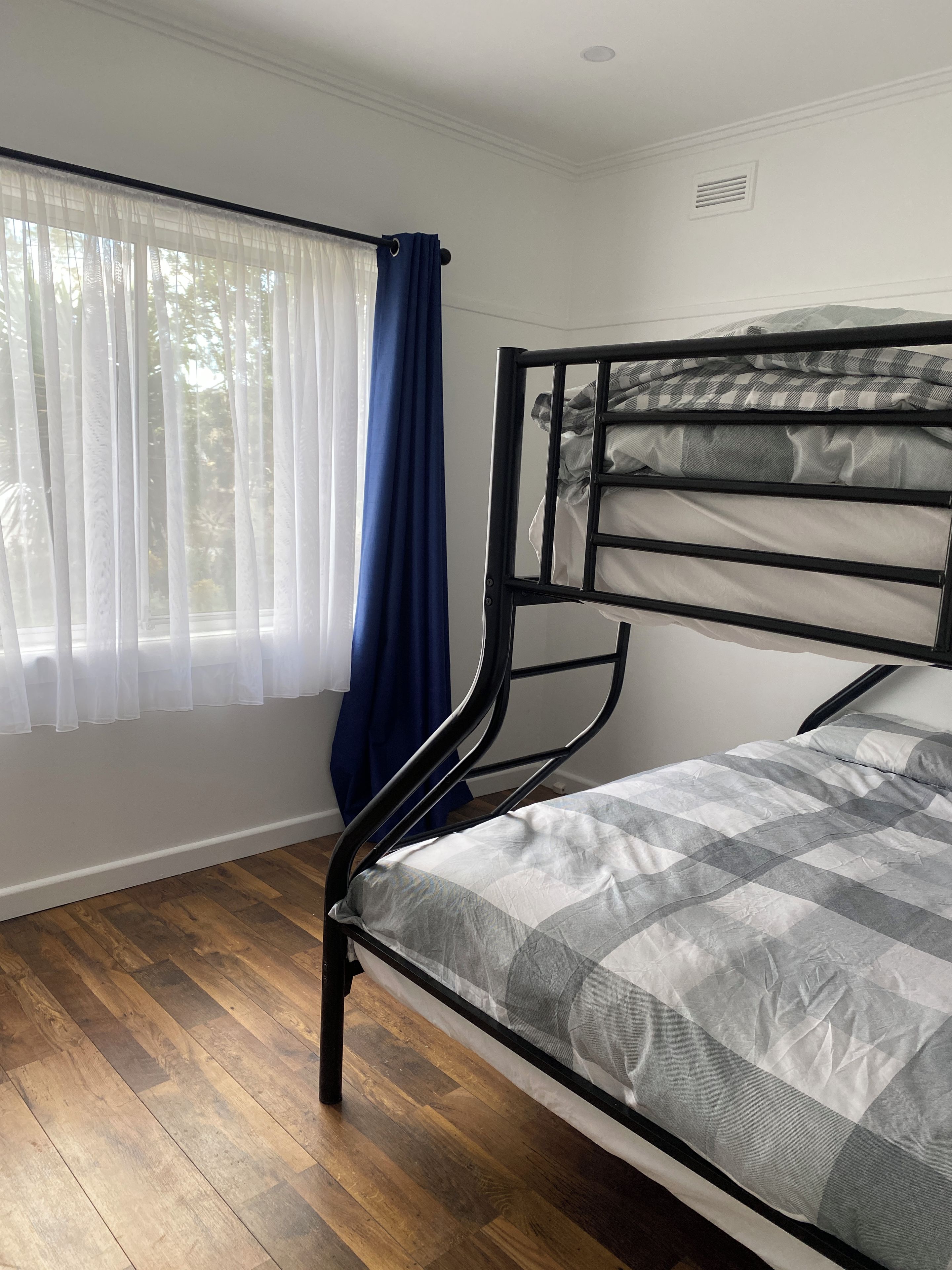 Una habitación con literas, piso de madera, una ventana con cortinas traslúcidas y un respiradero a la vista.