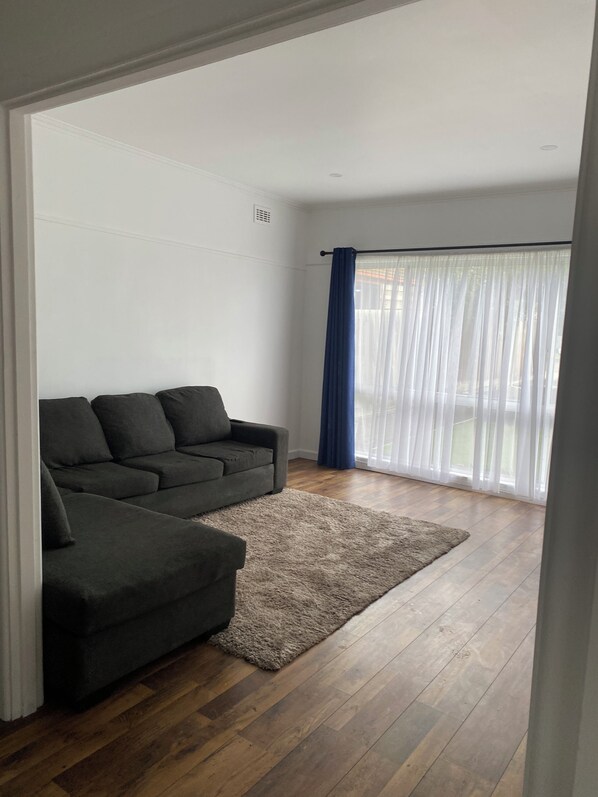 A living room with a sectional sofa, a rug, wooden flooring, and a large window with sheer curtains.