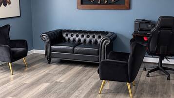 A room with a blue wall, a black leather sofa, two black chairs with gold legs, and a framed artwork.