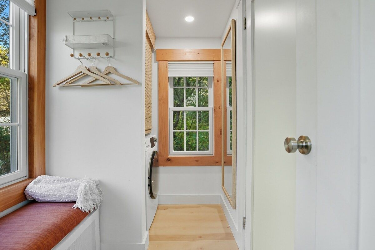 A narrow hallway with a wooden bench, a window with a view of greenery, and a door with a mirror.