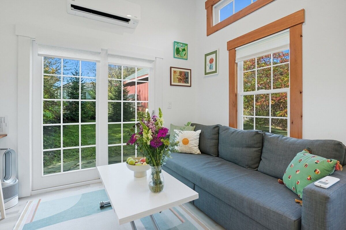 A living room with a grey sofa, a white coffee table, and a vase of flowers. There are two windows with wooden frames and a view of greenery outside.