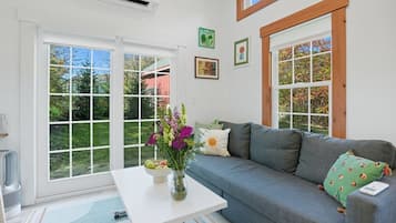 A living room with a grey sofa, a white coffee table, and a vase of flowers. There are two windows with wooden frames and a view of greenery outside.