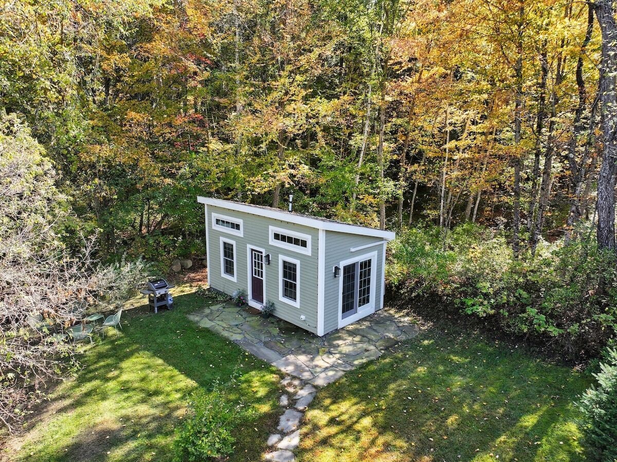 A small, single-story house with a stone pathway leading to the front door, surrounded by dense trees and a grassy yard.