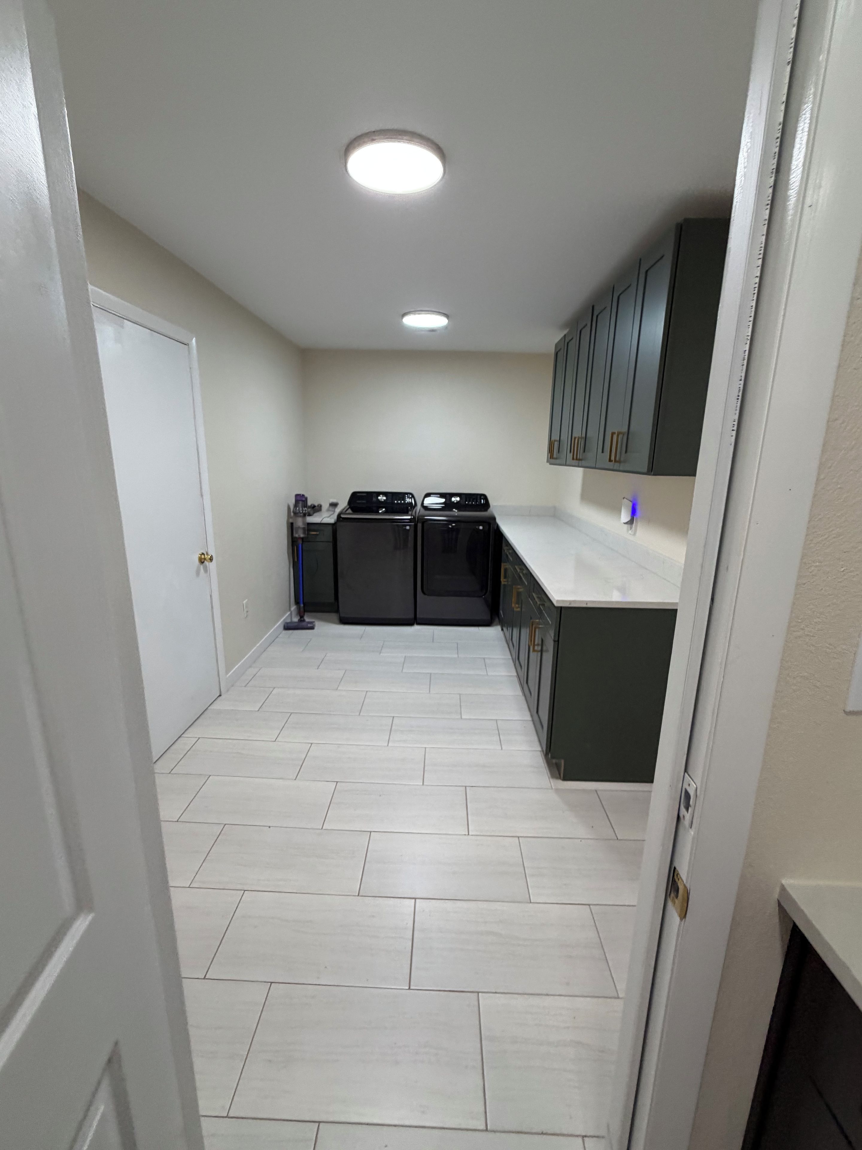 A narrow laundry room with a white door, two stacked washers and dryers, a mop, and a white tiled floor.