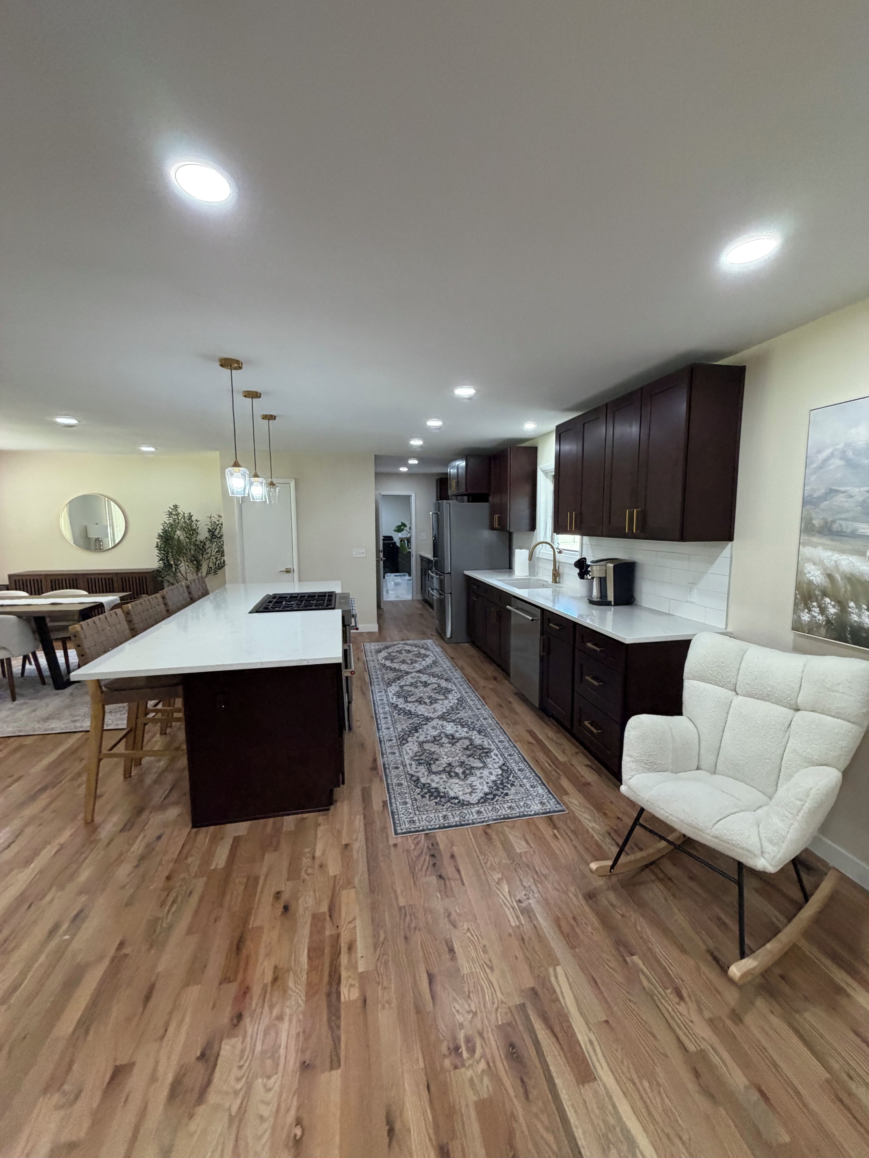 A modern kitchen with wooden flooring, a central island, dark cabinets, and recessed lighting.