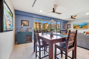 A dining area with a wooden table and chairs, a blue cabinet with wine bottles, and a view of buildings through large windows. - Medano Beach, Walk Everywhere, Pool, Jacuzzi, 2BD (Cabo San Lucas)
