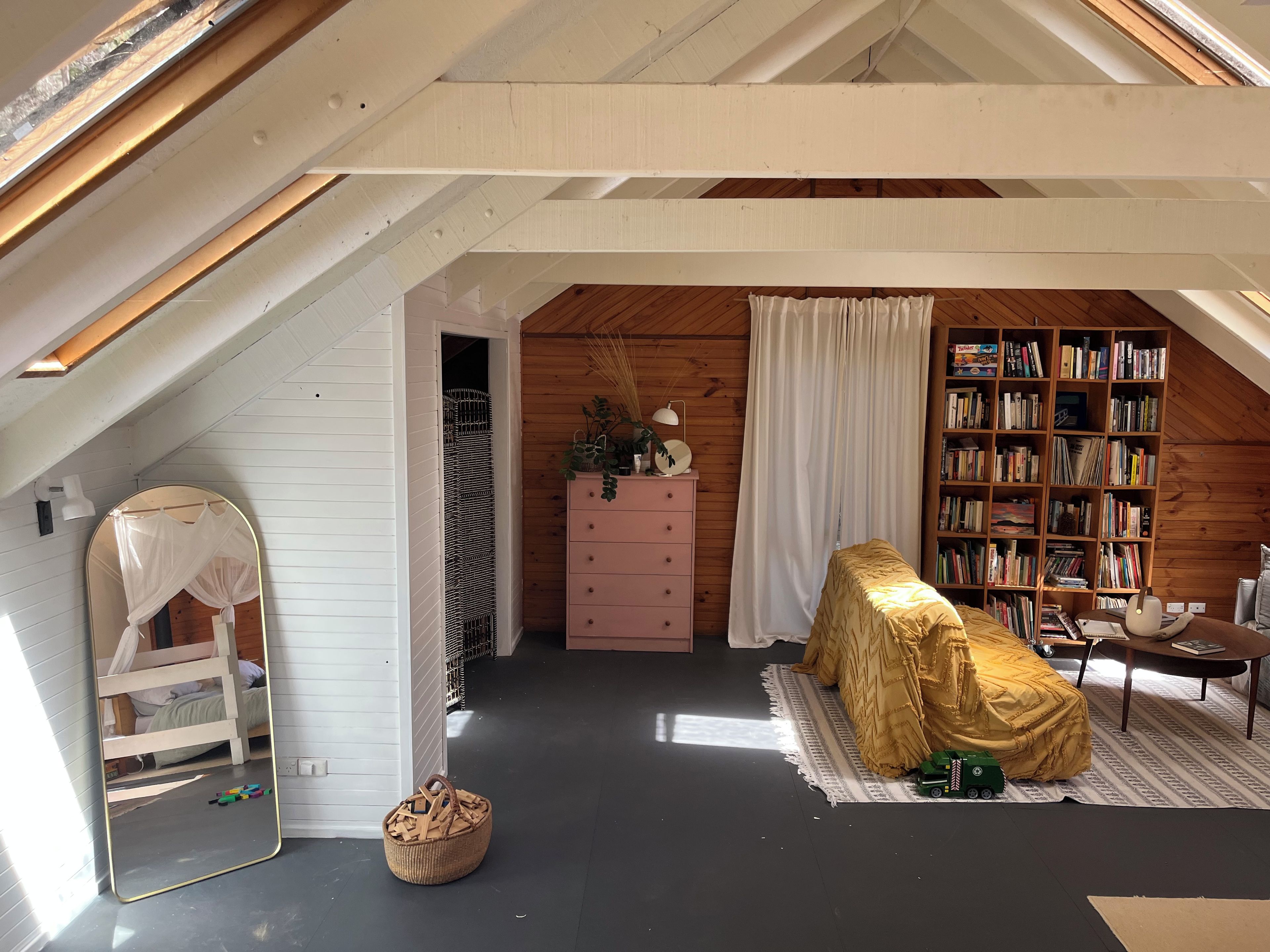 A cozy attic bedroom with a sloped ceiling, a large mirror, a pink dresser, a bookshelf, and a covered chair.