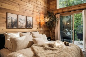 A wooden interior with a bed, pillows, a chandelier, and a view through a glass door. - Waterfront King Suite | Hot Tub | Forest Views | Romantic Getaway (Sevierville)