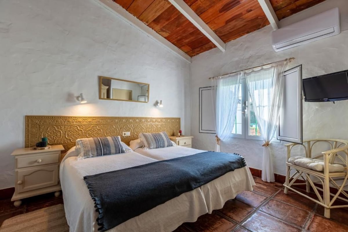 Quarto com duas camas, teto de madeira, televisão, janela com cortinas e uma cadeira.
