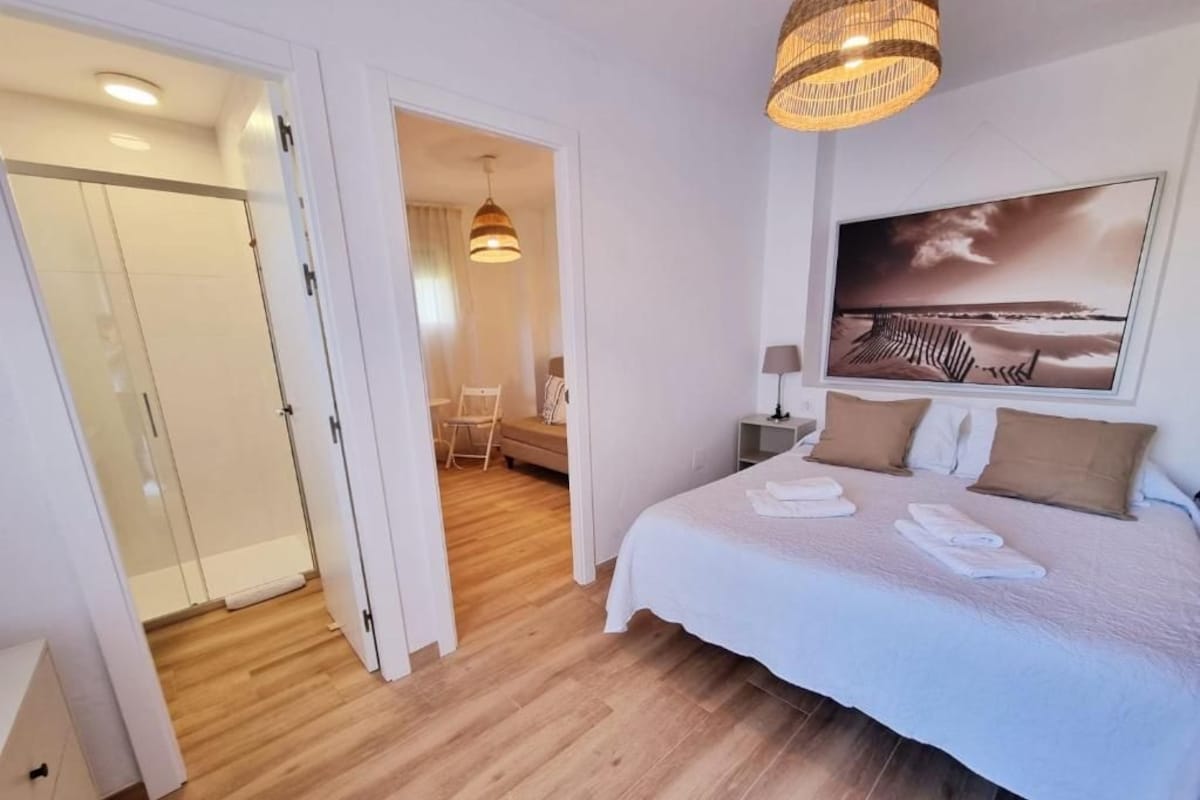 Quarto de hotel com uma cama, um quadro, um abajur e uma porta que dá para outro espaço.