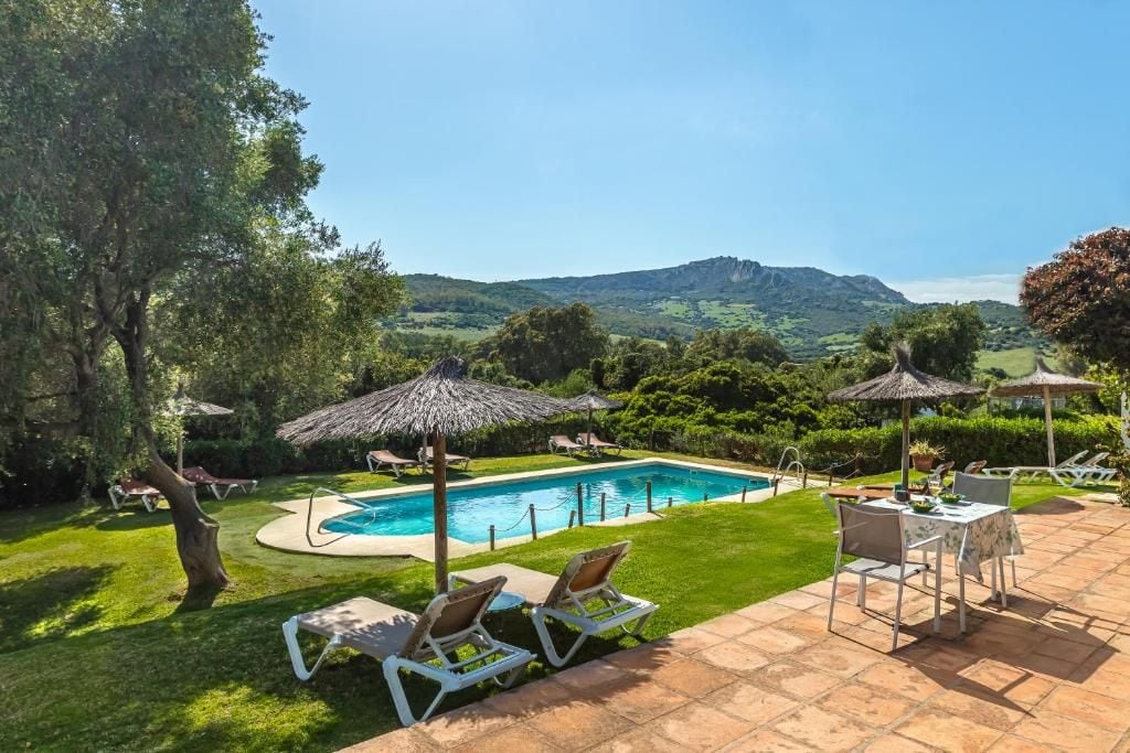 A swimming pool with lounge chairs and umbrellas, surrounded by a well-maintained lawn and a view of distant hills.