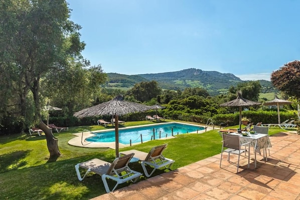 Piscina com espreguiçadeiras e guarda-sóis, cercada por um gramado bem cuidado e vista para colinas ao longe.
