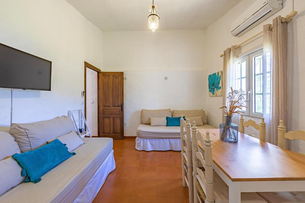Una habitación con cama, sofá, mesa de comedor y una silla. Tiene una puerta, una ventana con cortinas y un aire acondicionado de pared.