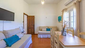 Una habitación con cama, sofá, mesa de comedor y una silla. Tiene una puerta, una ventana con cortinas y un aire acondicionado de pared.