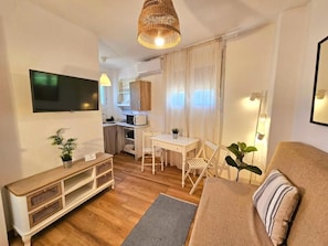 Um espaço de estar compacto com sofá, mesa de jantar, TV fixada na parede e uma cozinha compacta ao fundo.