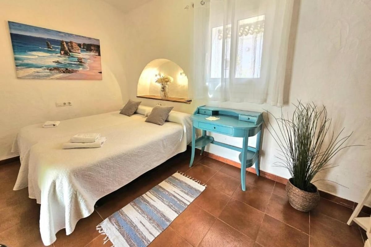 Quarto com cama, escrivaninha, planta em vaso e um quadro na parede.