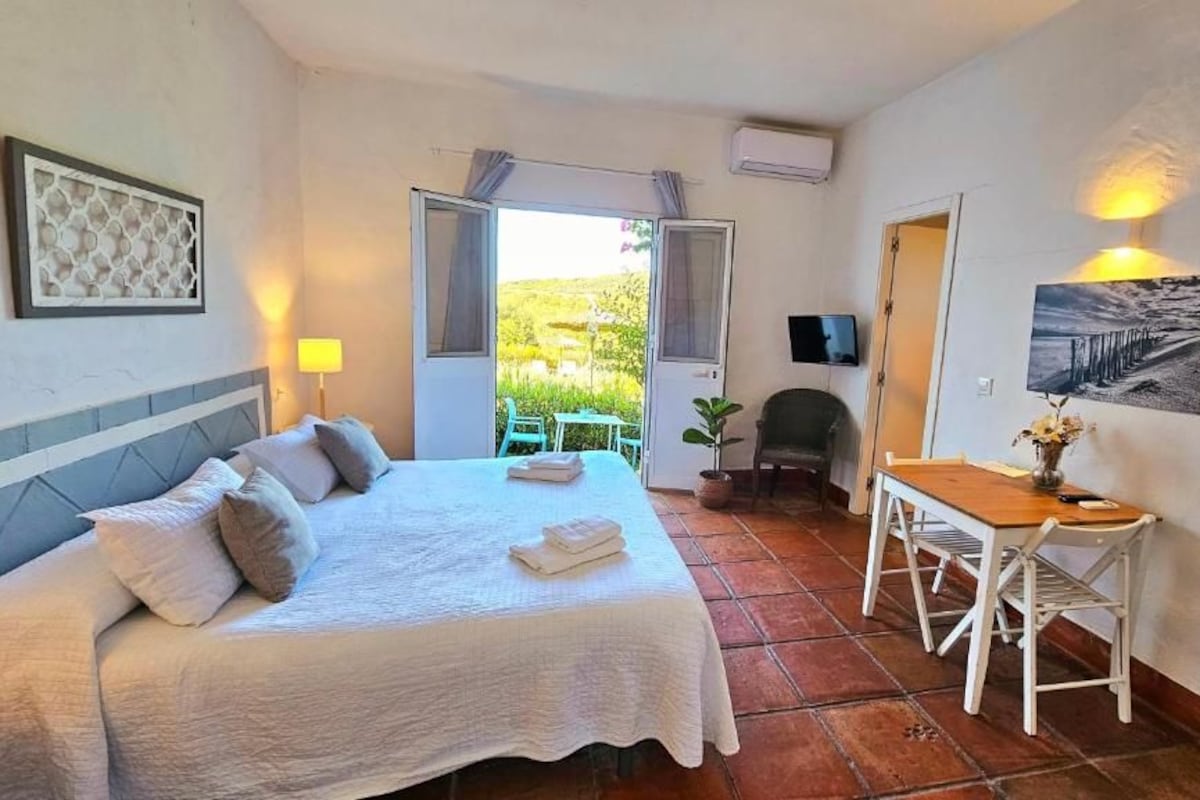 Quarto de hotel com cama, escrivaninha, cadeira, televisão e vista para o exterior.