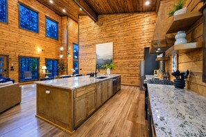 A spacious kitchen with wooden walls, a large island, and a view of the outdoors through the windows. - Broken Bow Bliss · Private Heated Pool, Hot Tub & Arcade Game Room | Sleeps 12 (Broken Bow)